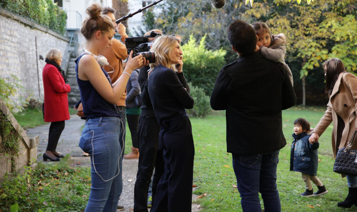 Equipe de tournage dans un jardin. Dans les bras de l'auteur de dos, une petite fille. Sur la gauche, la productrice les regarde en souriant.