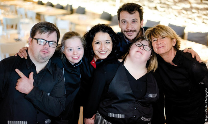 Photo de l'équipe Le Reflet, réunissant personnes atteintes de trisomie et "valides", vue de face dans la salle du restaurant. Habillés en noir, ils sourient.