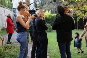 Equipe de tournage dans un jardin. Dans les bras de l'auteur de dos, une petite fille. Sur la gauche, la productrice les regarde en souriant.