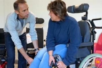 Un homme est penché vers une femme en fauteuil roulant. Il saisit quelque chose sur le côté du fauteuil en lui parlant. Les deux se regardent. 