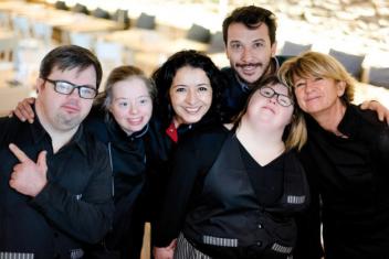 Photo de l'équipe Le Reflet, réunissant personnes atteintes de trisomie et "valides", vue de face dans la salle du restaurant. Habillés en noir, ils sourient.