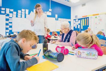 Une salle de classe aux murs bleus. Au premier plan, un table carré de quatre places. De chaque côté deux personnes sont assises : deux enfants d'une huitaine d'années d'un côté, une petite fille et une dale âgée de l'autre. Tous écrivent. Des troussent d'écoliers reposent sur la table. Au second plan, debout devant la table, l'institutrice tient une feuille à la mai et s'adresse à la dame âgée 
