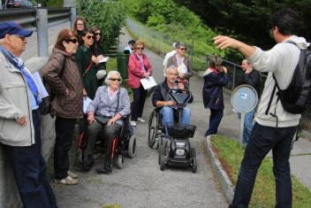 A l'exterieur, au bord d'une route, sur un chemin pieton dans ce qui semble être un parc un homme de dos parle à un groupe aligné devant lui. Dans ce groupe : deux personnes en fauteuil roulant, des seniors et des plus jeunes. 