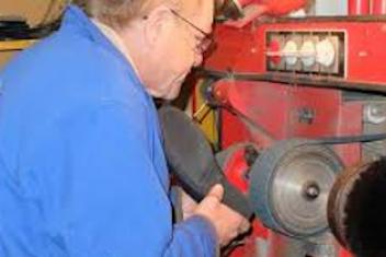Cordonier en train de travailler. Il porte une veste bleu et est en train de polir une chaussure avec une machine spécialisée. 