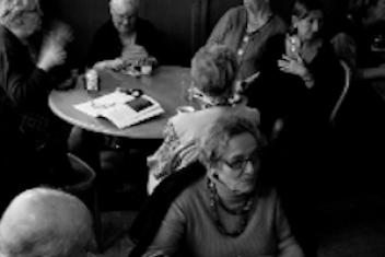 Photo en noir et blanc. Des personnes âgées sont assises autour de table de bistrot et discutent entre elles