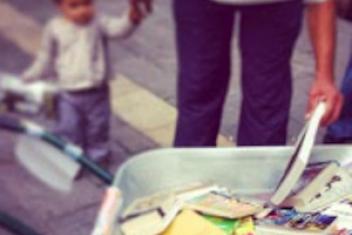 Un homme tient par la main un très jeune enfant et est penché au dessus d'une brouette dans laquelle se trouve plein de livres. Ils les regardent et en tient un dans sa main