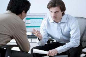 Deux hommes sont assis en face l'un de l'autre, en train de discuter. L'homme face à la caméra a un papier roulé dans sa main gauche.