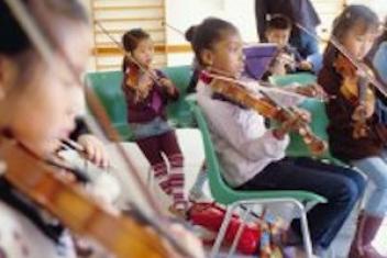 Des jeunes filles, et jeunes enfants sont en train de jouer de la musique. L'une est au premier plan, floutée, et trois autres enfants sont dans l'arrière plan. Tous ont un violon dans les mains