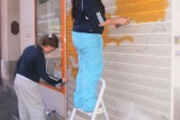 Deux jeunes filles sont en train de repeindre un rideau de fer en orange. L'une est sur un escabot et l'autre est debout, sur l'arrière plan de l'image 