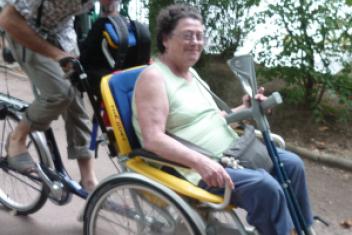 Un jeune homme est à vélo et sur l'avant du vélo se trouve un fauteuil dans lequel est installée une femme, béquilles en mains. Les deux personnes souris à la caméra. 