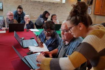 AU premier plan, une fille debout à coté d'un senior pointe du doigt vers un ordinateur portable placé sur la table devant lui. A coté de lui, une dame regarde également l'écran. Au fond, au second plan, autour de la même grande table deux seniors, une femme et un homme, sont assis devant un ordinateur. Debout entre les deux, une jeune fille est penchée vers l'ordinateur. Derrière eux, de dos, un autre couple jeune fille / personne agée est également devant un ordinateur.